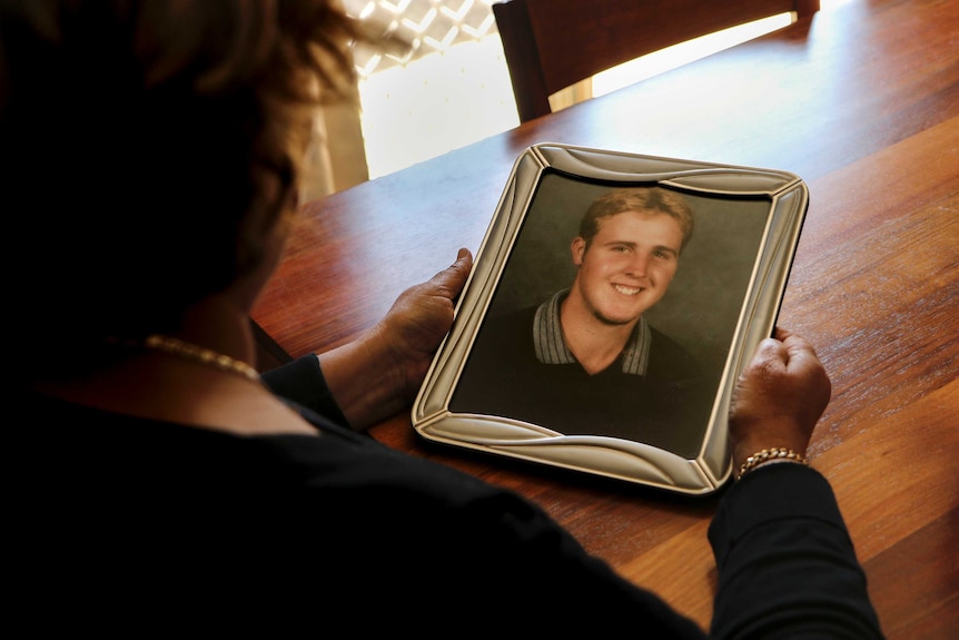 Woman looks at picture frame with a photo of her son, sitting at wooden table.
