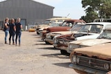 Three women on left looking at line up of rusty cars on right
