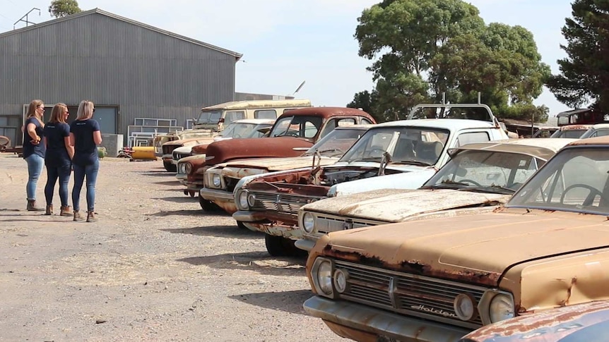 Three women on left looking at line up of rusty cars on right