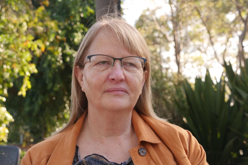 A bespectacled woman with shoulder-length hair stands outside, looking solemn.