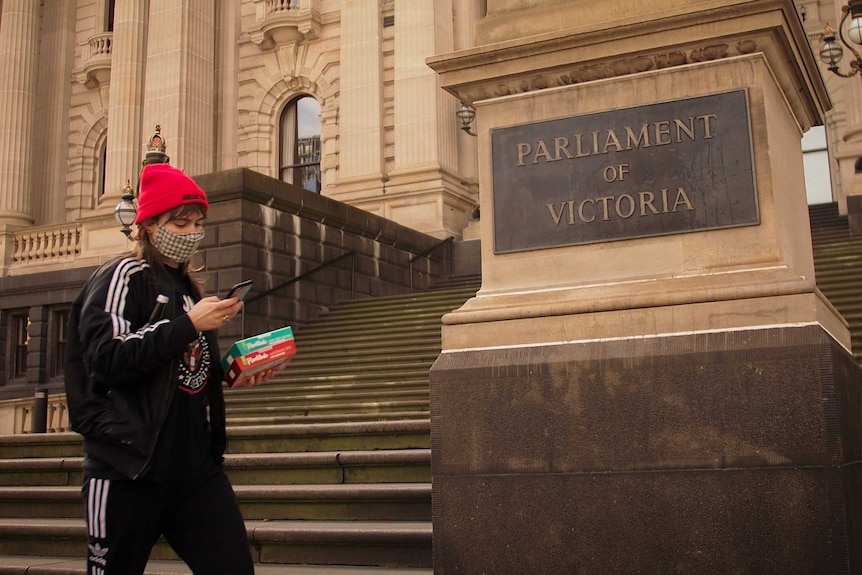 A woman in a red beanie wearing a mask looks at her phone as she walks past a Victorian Parliament sign.
