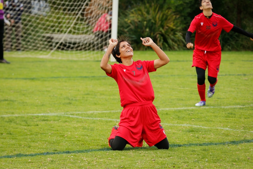 A footballer wearing a red uniform is on her knees, arms raised to the sky, celebrating a goal.