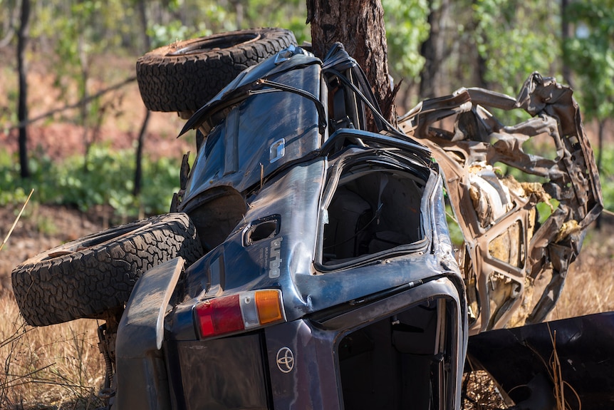 A blue landcruiser lies on its side against a tree. It is extensively damaged. 