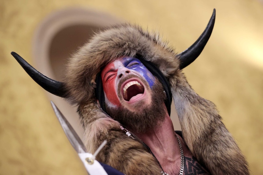A man wearing a horned fur hat holding a spear yells inside the US Capitol Building while holding a spear.