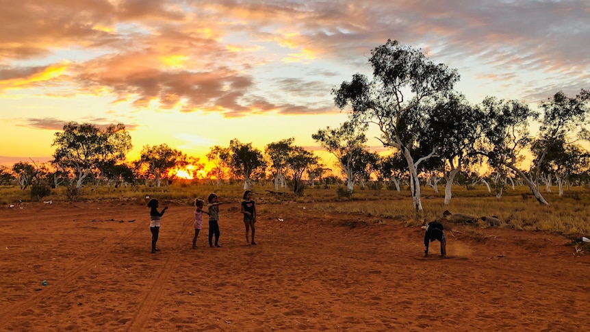 A group of children play in the red dirt as a sun sets behind them