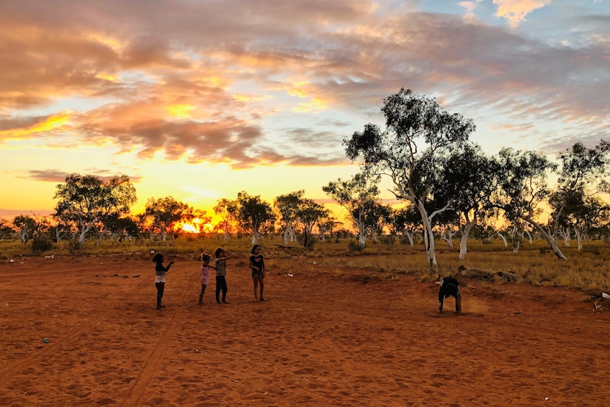 A group of children play in the red dirt as a sun sets behind them