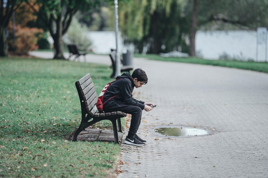 A young boy scrolling on this phone on a bench in a park. 