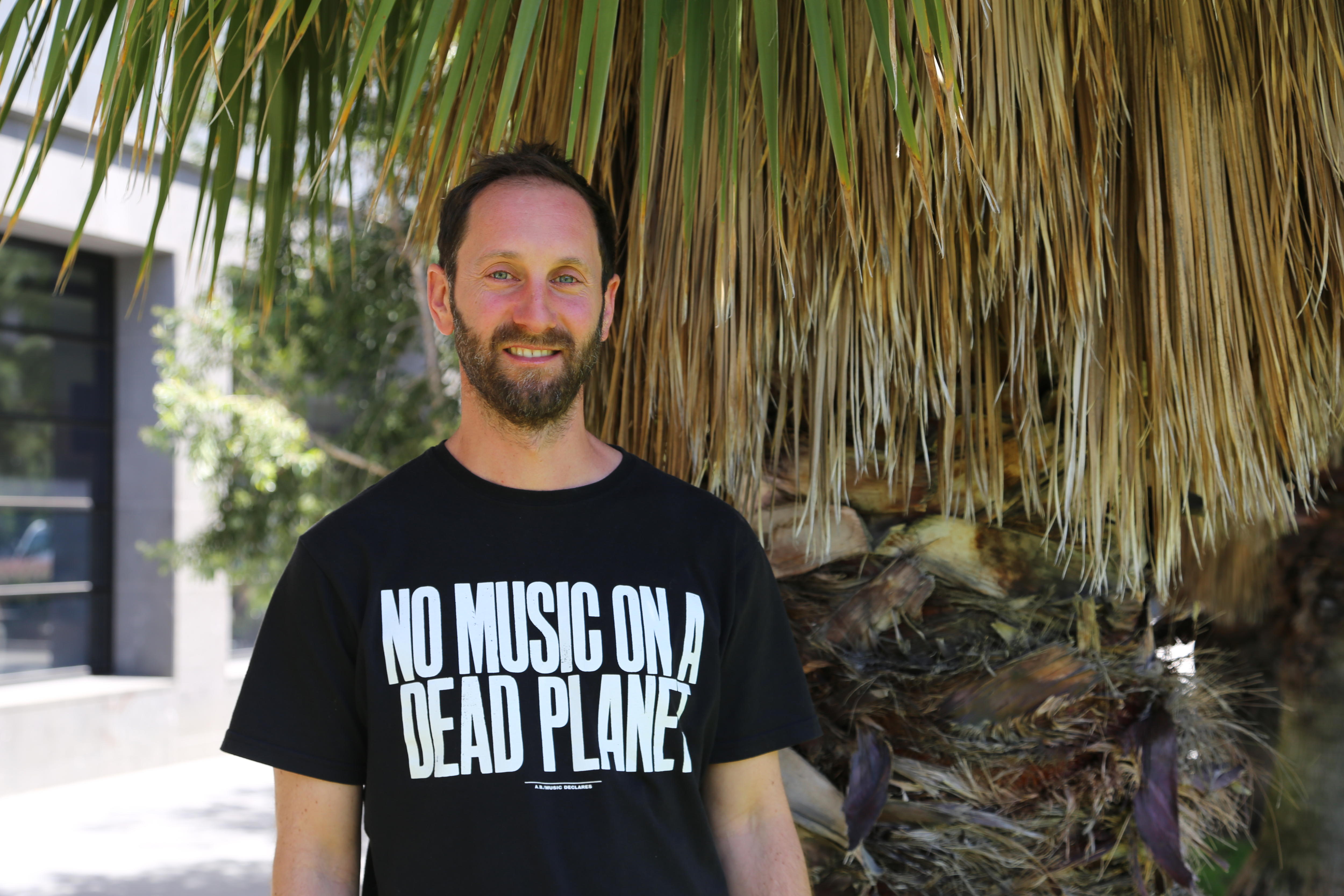 Un hombre con barba sonríe junto a un árbol. Su camisa negra dice: "No hay música en un planeta muerto.".