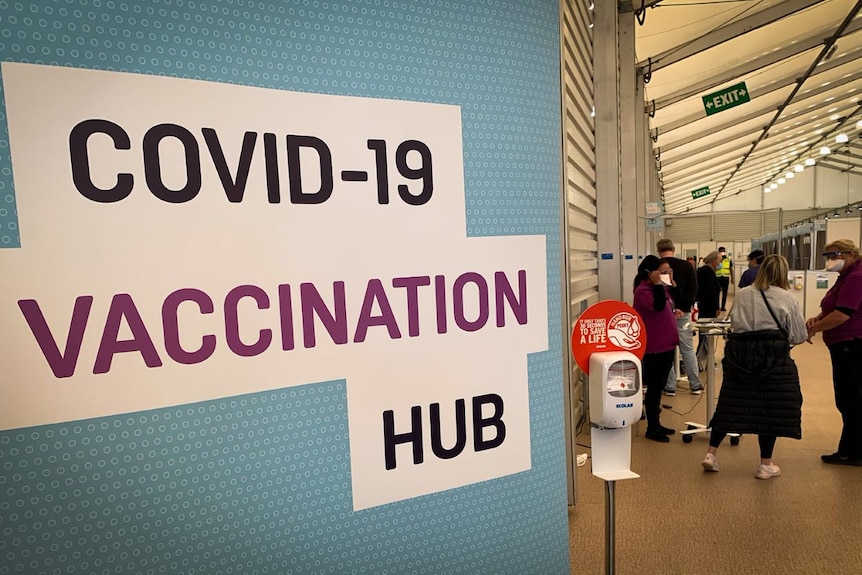 Unidentifiable people wait in a room with a sign saying it is a vaccination hub for COVID-19.