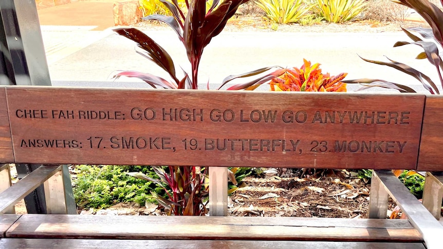 A park bench inscribed with words: Chee Fah riddles: Go High Go Low Go Anywhere; Answers: 17. Smoke; 19. Butterfly; 23: Monkey