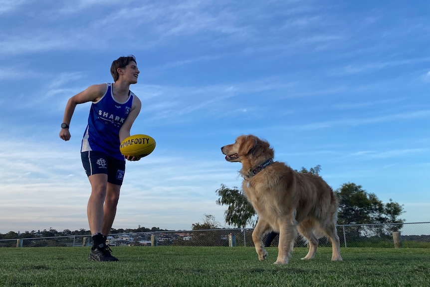 Footy player handballs a yellow footy while a golden retriever watches excitedly.