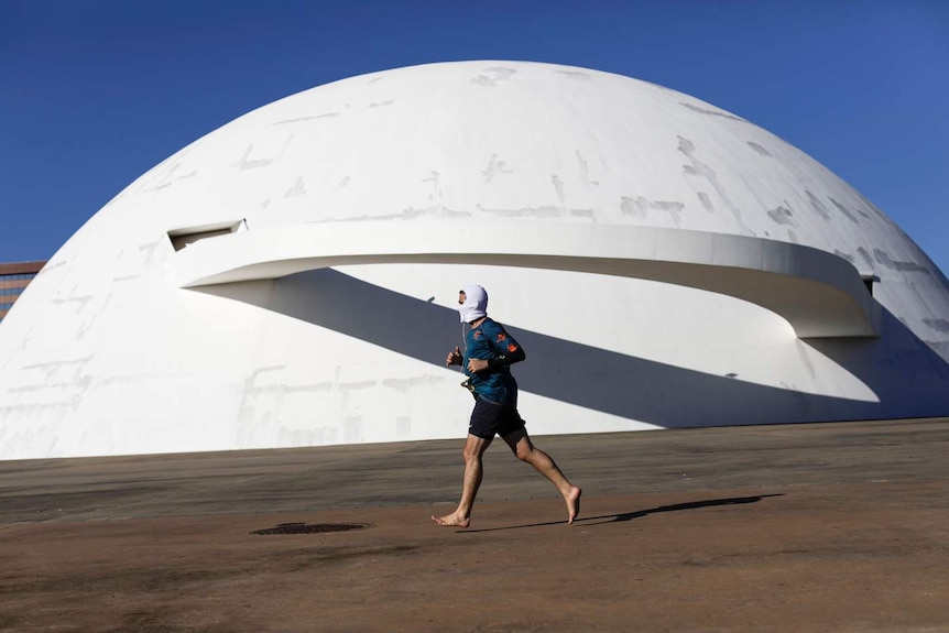 A man jogs wearing a white face mask in front of a white spherical building on a blue day.