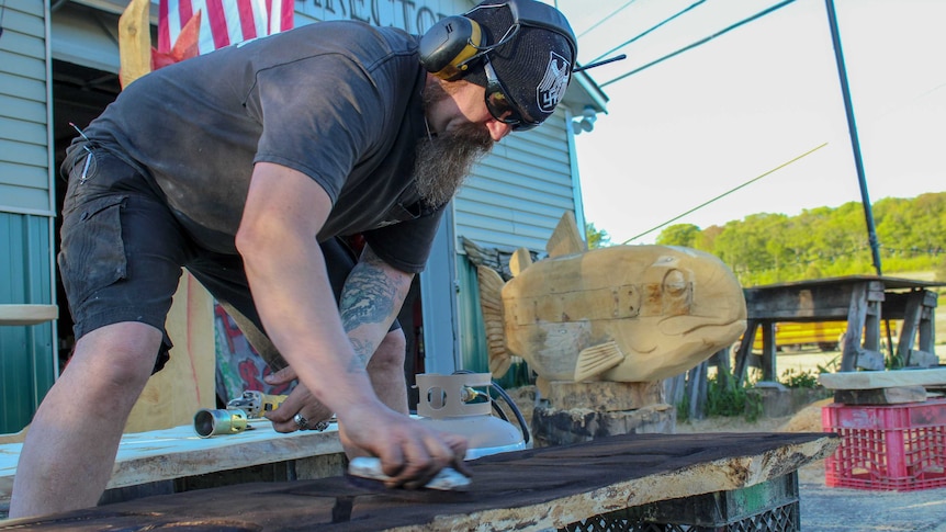 A man sands a piece of wood while wearing protective headphones