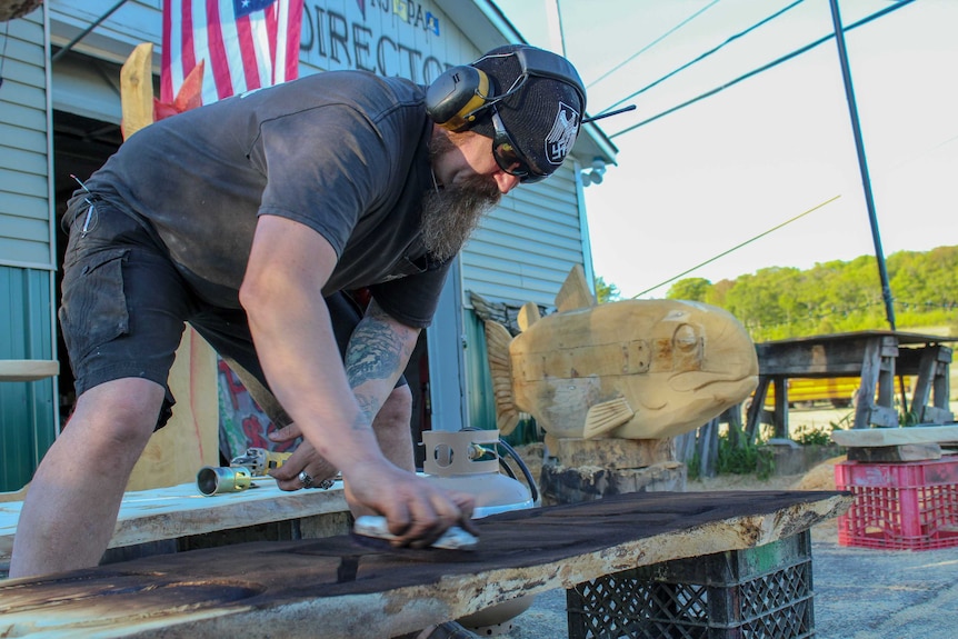 A man sands a piece of wood while wearing protective headphones