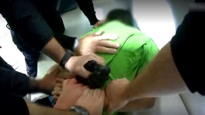 A boy wearing a green shirt lies face down. Several adults' hands hold him and his arms.