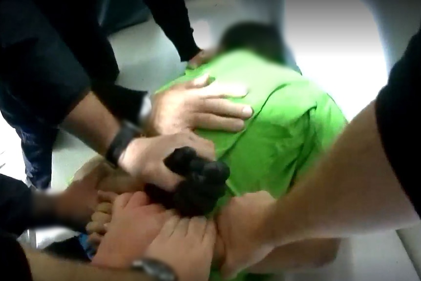 A boy wearing a green shirt lies face down. Several adults' hands hold him and his arms.