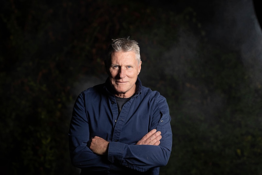 Michael Brissenden with arms crossed, smiling slightly, long-sleeved blue shirt, black background