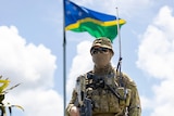 An ADF soldiers wearing a mask and sunglasses in uniform stands in front of a flag pole with the Solomon Islands flag