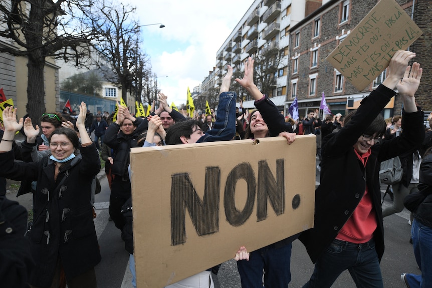 A group of protesters is pictured from the front, one of them holds a large cardboard sign that says 'NON'. 