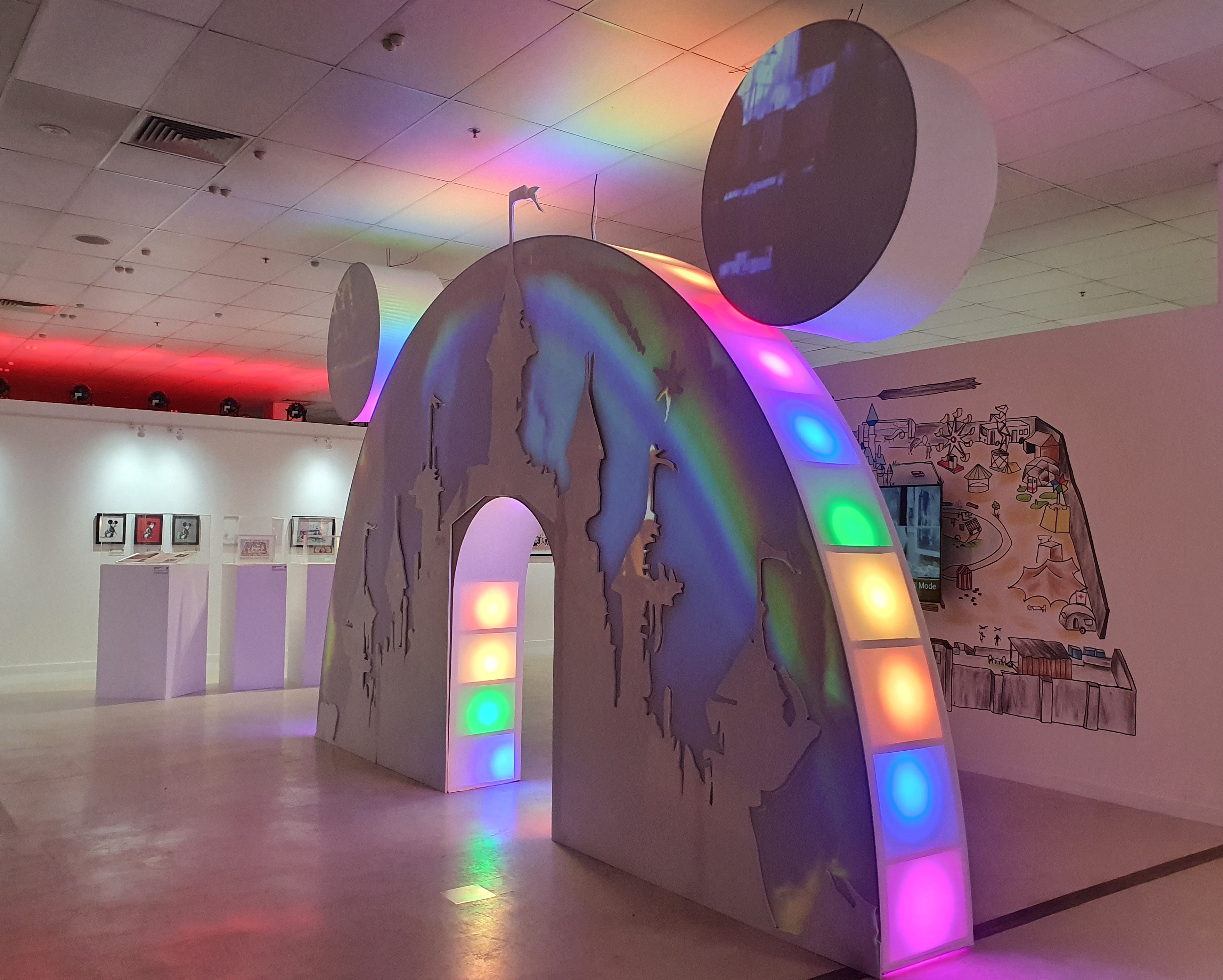 A doorway shaped like Mickey Mouse ears with bright colours projected onto it, seen at an exhibition.
