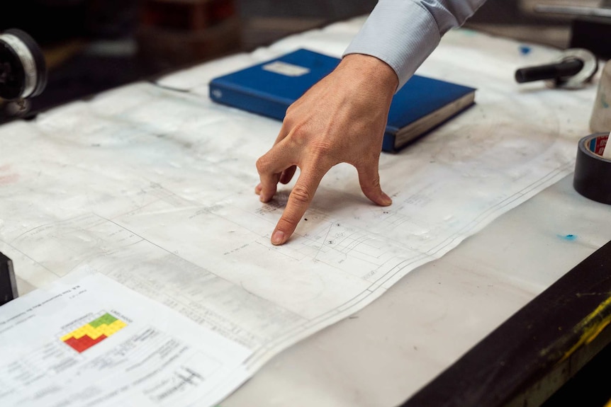 A close-up shot of a man's hand pointing at blueprints on a table.