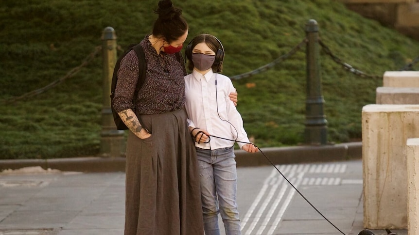 A woman hugs a young girl by the side of a road. The girl holds a dog lead, and both wear masks.