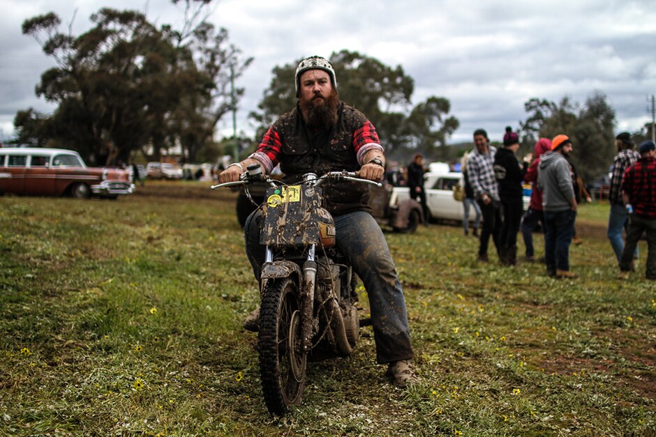 A man in an open-faced motorbike helmet sits on a muddy motorbike.