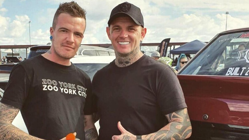 Two men with heavily tattooed arms standing next to one another and smiling