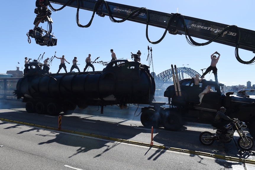 A large camera crane films people atop a big vehicle with the Sydney Harbour Bridge in the background.