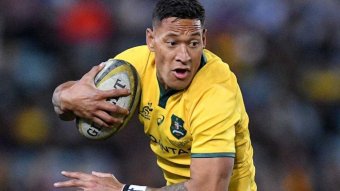 Un joueur de rugby vêtu de noir s'attaque à un joueur vêtu de jaune autour des hanches tandis qu'un autre joueur en noir regarde.