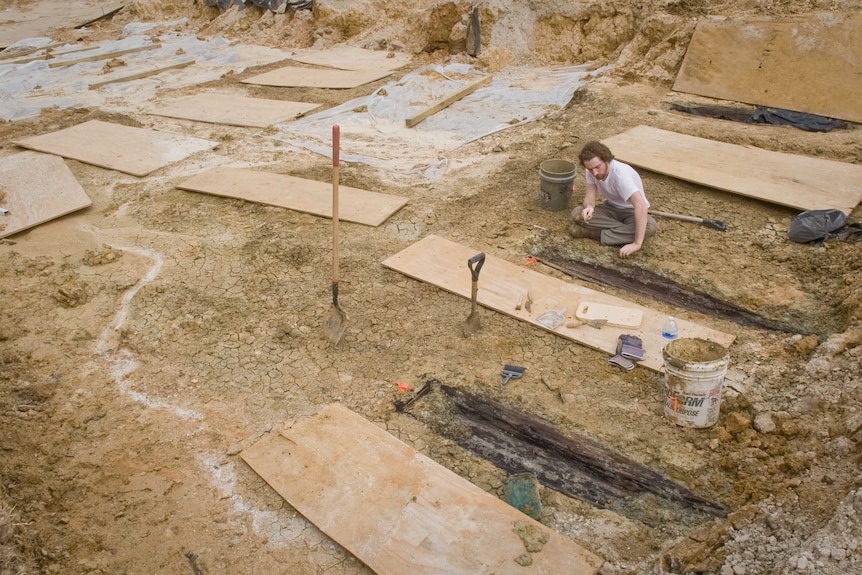 A man sits by a half buried coffin, surrounded by other grave sites.