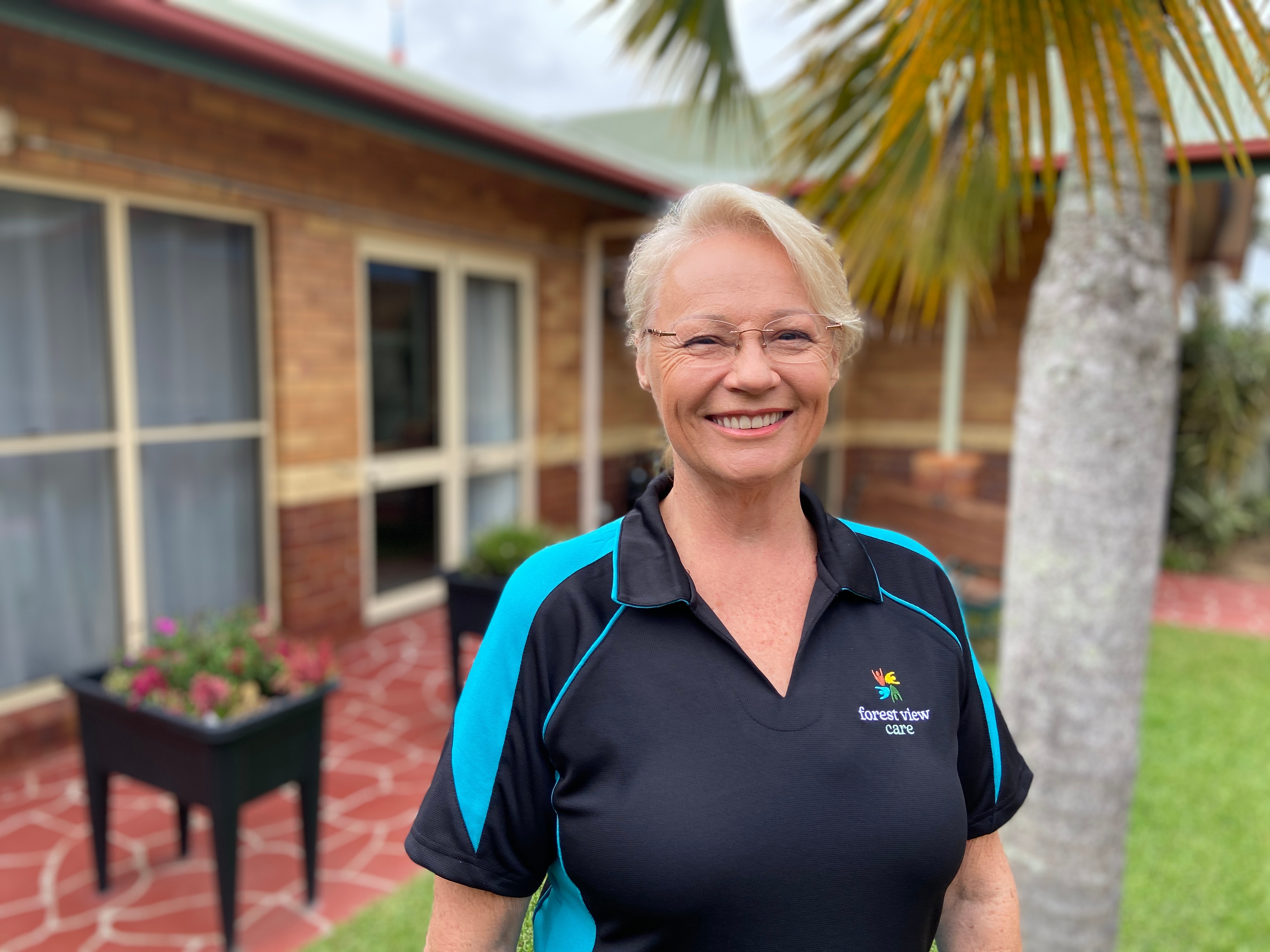 A middle-aged woman in a garden courtyard, smiling at the camera.