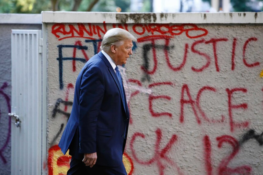 Donald trump walks past a wall on which 'no justice no peace' is scrawled amongst other graffiti.