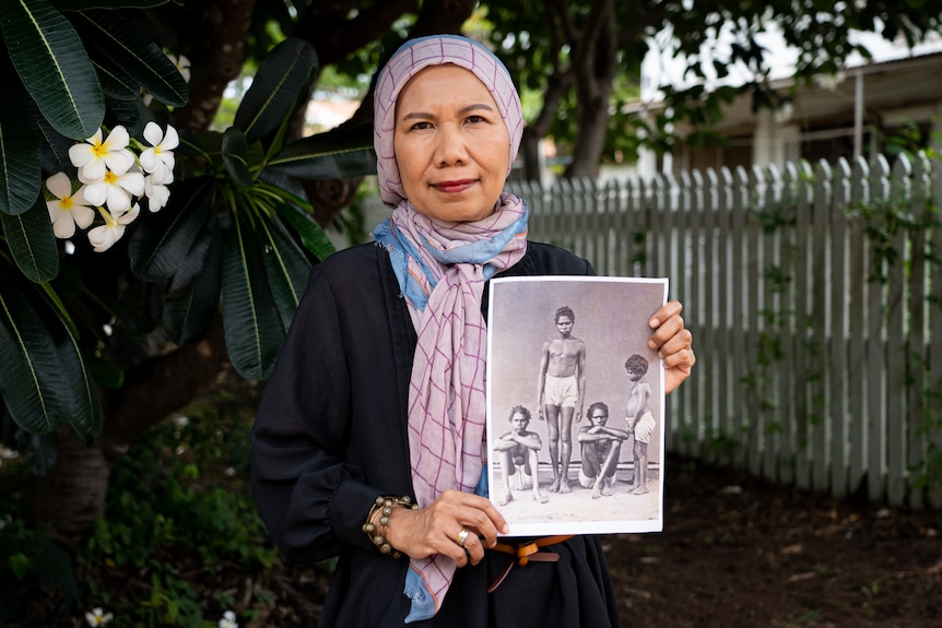 A woman stands in front of a tropical tree smiling and holding a black and white photograph