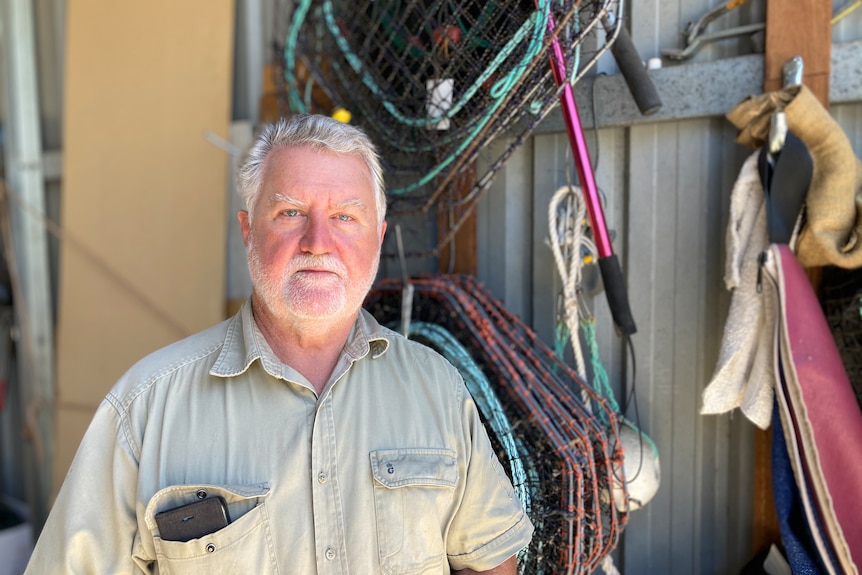 A man in a khaki shirt stands in front of a shed wall, there's a lot of fishing gear hanging behind him