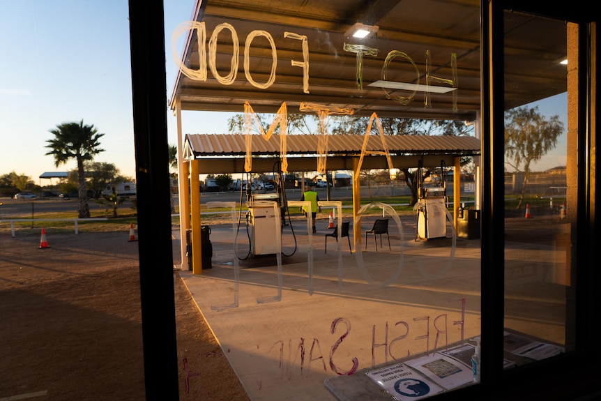 Fuel bowsers are seen through a window with colourful writing that says "hot food, atm, coffee" at sunrise.