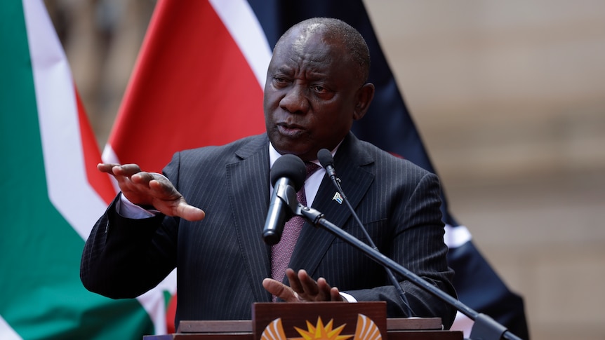 A dark-skinned man in a suit gestures at a lectern, with the South African flag drapped behind him.