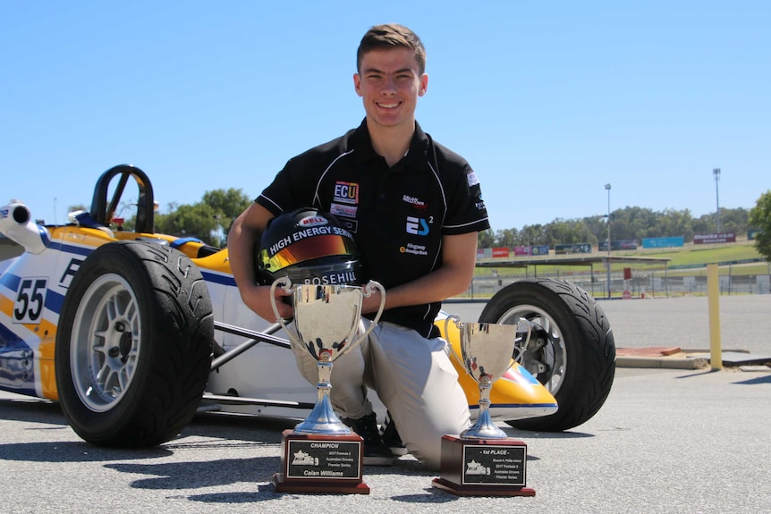 Calan Williams kneels in front of a white, blue and yellow racing car with sports trophies in front of him