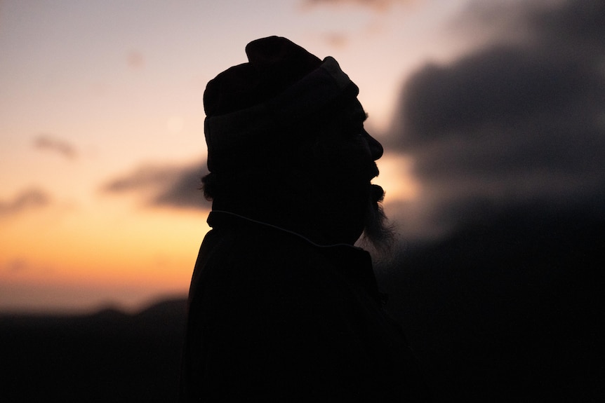 A silhouette of an older Aboriginal man with his mouth open. Clouds part in front of a golden sunrise over mountain ranges.