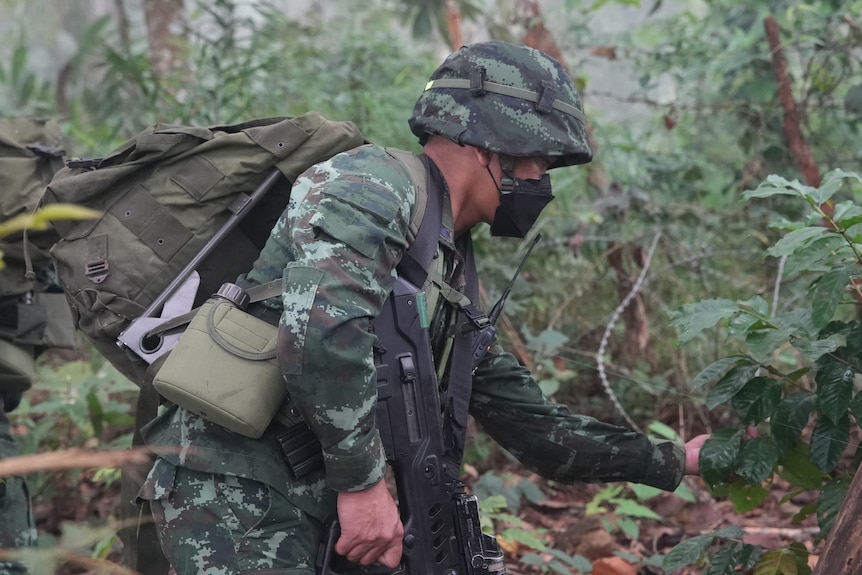 A soldier in camouflage gear carries a large backpack and gun. He leans over to check a sensor