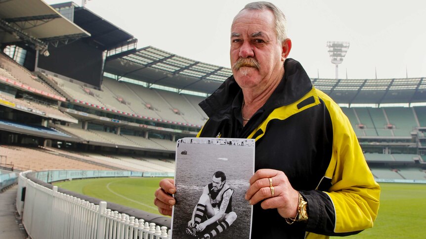 Former footballer Rober McGhie holds a picture of himself as a footballer, sitting on the ground smoking.