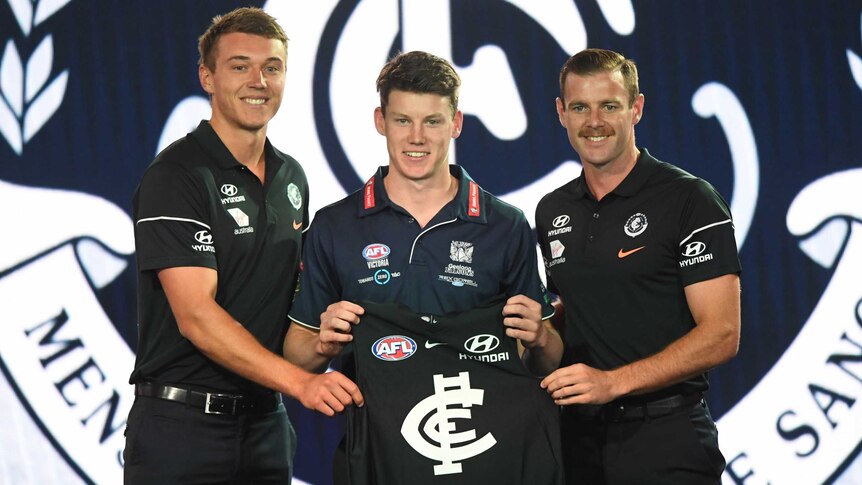 A prospective AFL footballer stands holding a Carlton jersey, with a Blues player and official at the 2018 national draft.