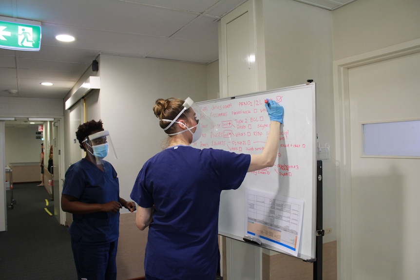 A nuse writes on a white board flanked by another nurse wearing full PPE