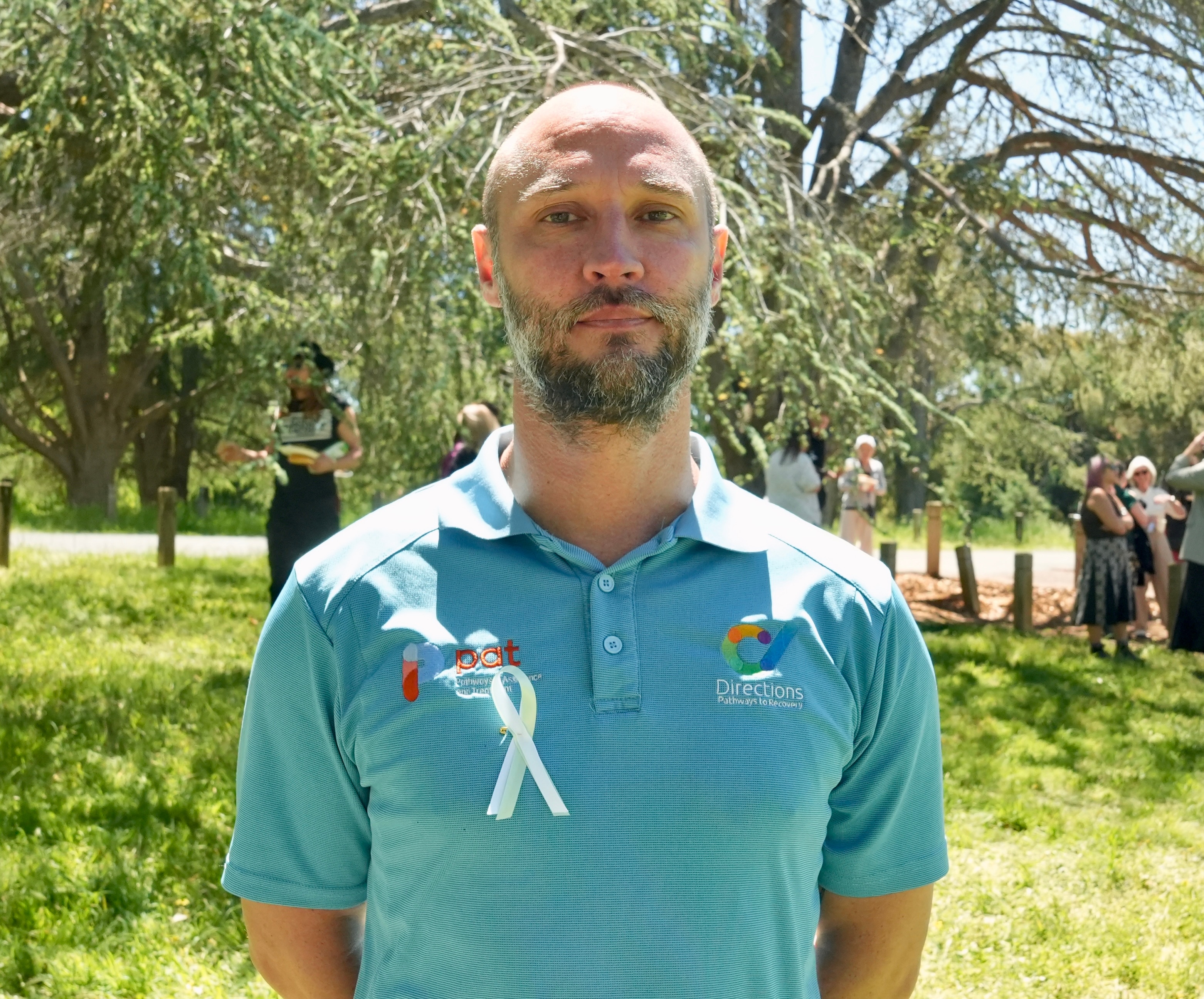 Man with a serious expression wearing a light blue collared t-shirt with a white ribbon pinned on it standing in a park