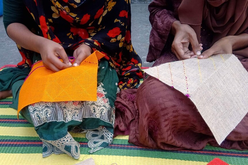 Two women sit hand sewing fabrics.