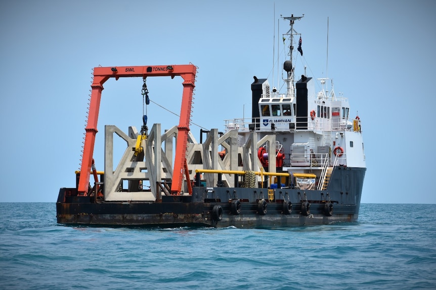 A 38m ship at sea carrying four concrete artificial reefs. They are a 3d  frame structure of a pyramid within a cube.