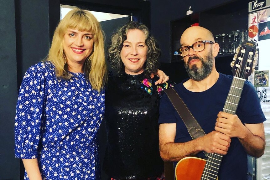 Sally Seltmann, Angie Hart and Simon Austin stand side by side. Simon holds a guitar, Sally's arm is around Angie's shoulders