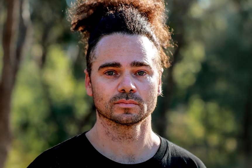 Man with black hair above head stands wearing black t-shirt amid trees and sunlight