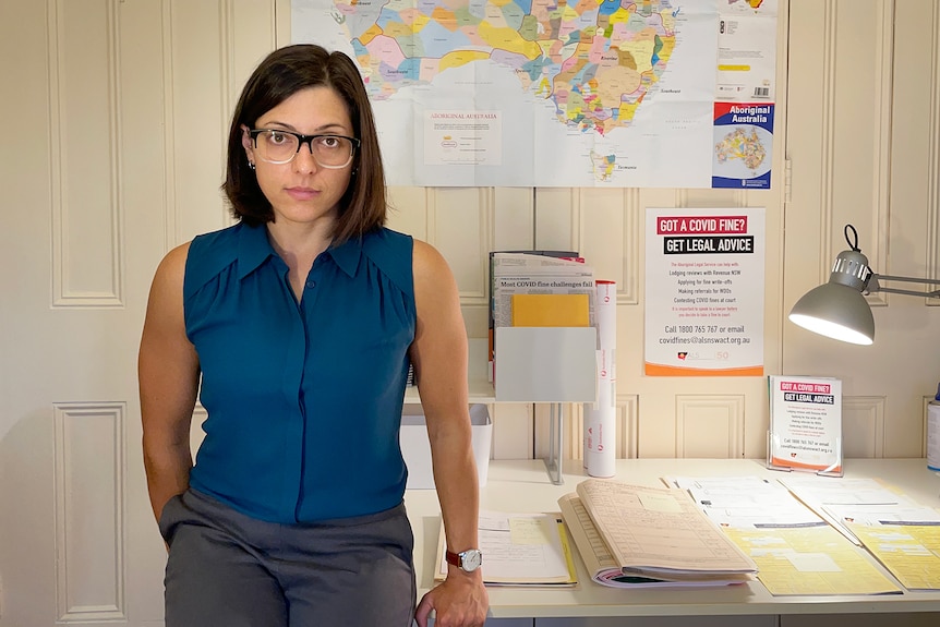 Woman with cropped dark hair and glasses in a dark blue short sleeved top, sitting on a desk, large map on wall behind