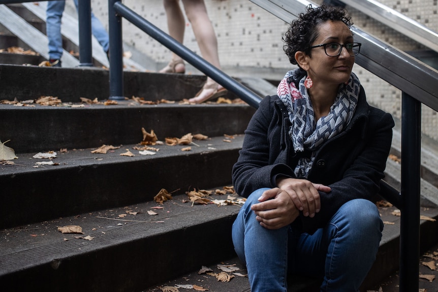 A woman with a colourful scarf and earrings sits on steps and looks away.